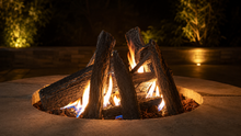 Load image into Gallery viewer, A circular outdoor fire pit with stacked logs and glowing orange-yellow flames, set in a stone burner at night with warm background lighting.