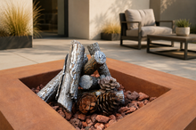 Load image into Gallery viewer, A square rust-orange outdoor fire pit with faux logs and pinecones inside, set on a patio with a blurred lounge chair and plants in the background.
