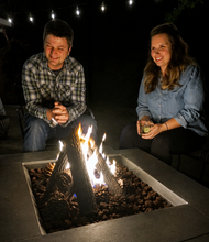 Load image into Gallery viewer, A man and a woman sitting beside a modern square fire pit table at night, with flames burning inside filled with dark stones. The man wears a plaid button-up shirt, and the woman wears a light blue denim shirt.
